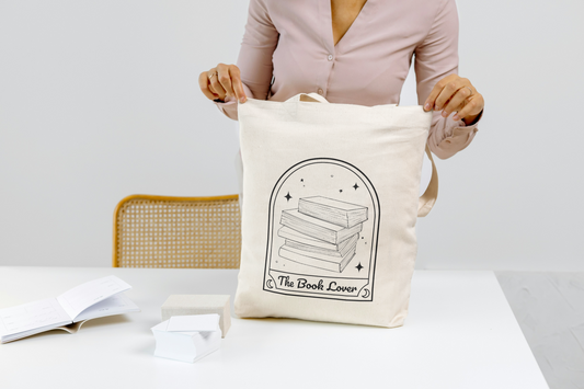 A person of colour is wearing a pale pinkshirt and holding a tote bag with a design that reads "The Book Lover" and has an illustration of a stack of books and sparkly stars all inside an arch with moon symbols on the corners.